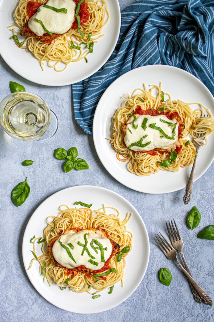 Overhead view of three white plate full of spaghetti and topped with Oven Baked Chicken Parmesan showing off melted mozzarella and a fresh basil garnish.