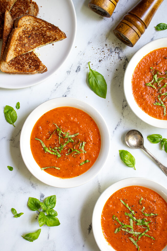 Overhead view of three bowls of roasted red pepper and tomato soup surrounded and garnished by fresh basil leaves.