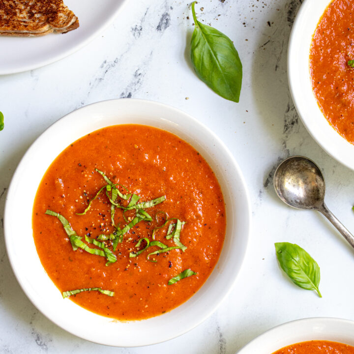 Close up of three bowls of tomato and red pepper soup on a countertop with basil leaves and spoon.