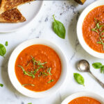 Pinterest Pin with text 'Roasted Red Pepper & Tomato Soup' image of three white bowls of tomato soup with basil leaves.