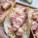 Pinterest Pin with text overlay 'Tuna Steak Tacos with sriracha mayo & asian slaw' images of a tray full of seared tuna tacos with garnishes.