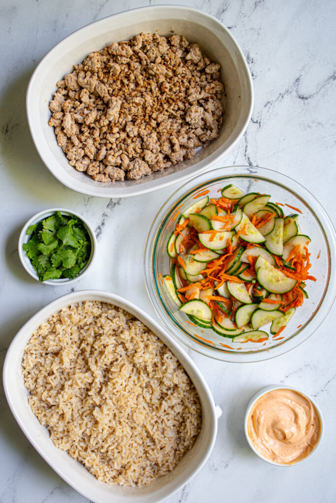 Different containers holding the different parts of the Banh Mi bowls, ground pork, brown rice, quick pickled veggies, sriracha mayo, and cilantro.