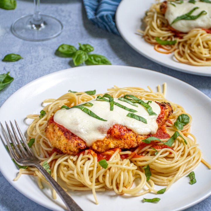 White plate piled high with pasta topped with an Oven Baked Chicken Parmesan covered in melted cheese and fresh basil garnish.