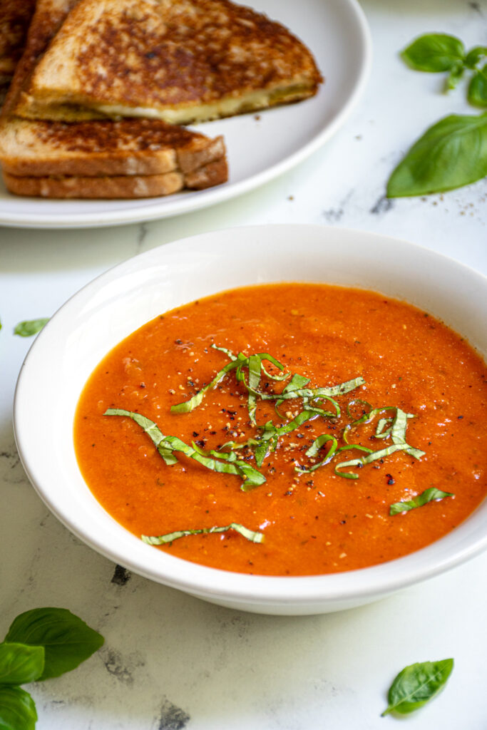 White bowl filled with roasted red pepper and tomato soup topped with freshly cracked black pepper and fresh basil leaves sitting next to a plate of grilled cheese.