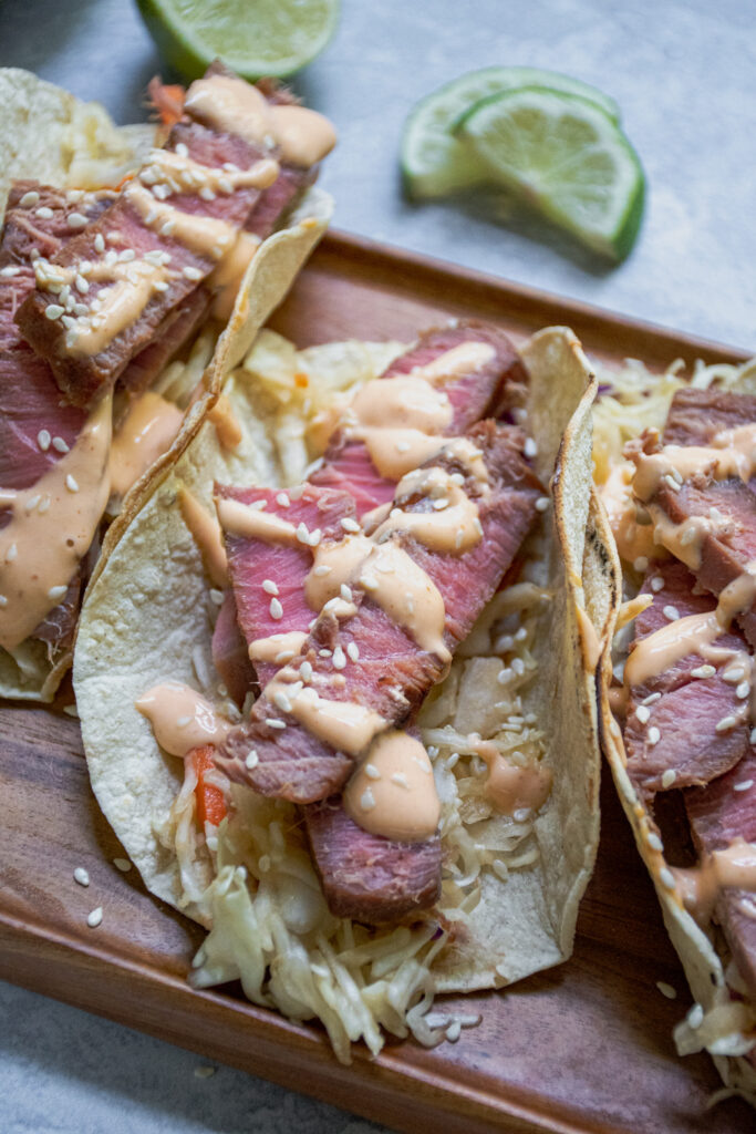 A side view of a close up of a tuna steak taco showing of slice tuna with pink middle garnished with sriracha mayo and sesame seeds.