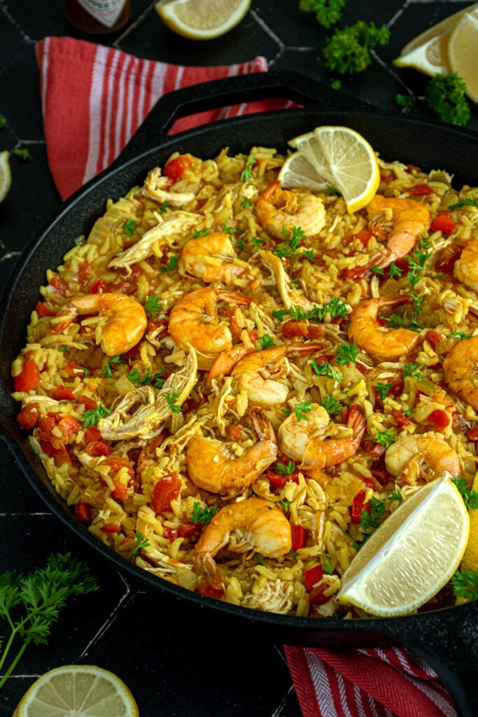 Angled view of a pan full of chicken shrimp paella showing off yellow rice, Reb bell peppers, chicken, and shrimp garnished with fresh parsley.