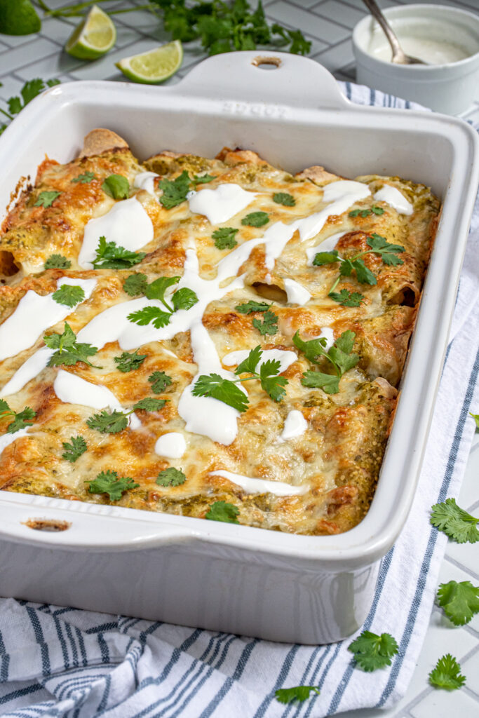A white baking dish filled with baked green chicken enchiladas topped with melty cheese, sour cream, and cilantro.
