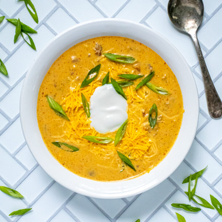 A white bowl on a white background filled with creamy yellow crockpot pumpkin chili topped with green onions and sour cream.