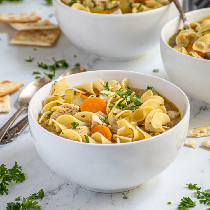A centered white bowl showing off a large portion of easy to make stovetop chicken noodle soup garnished with fresh parsley and served with saltine crackers on the side.