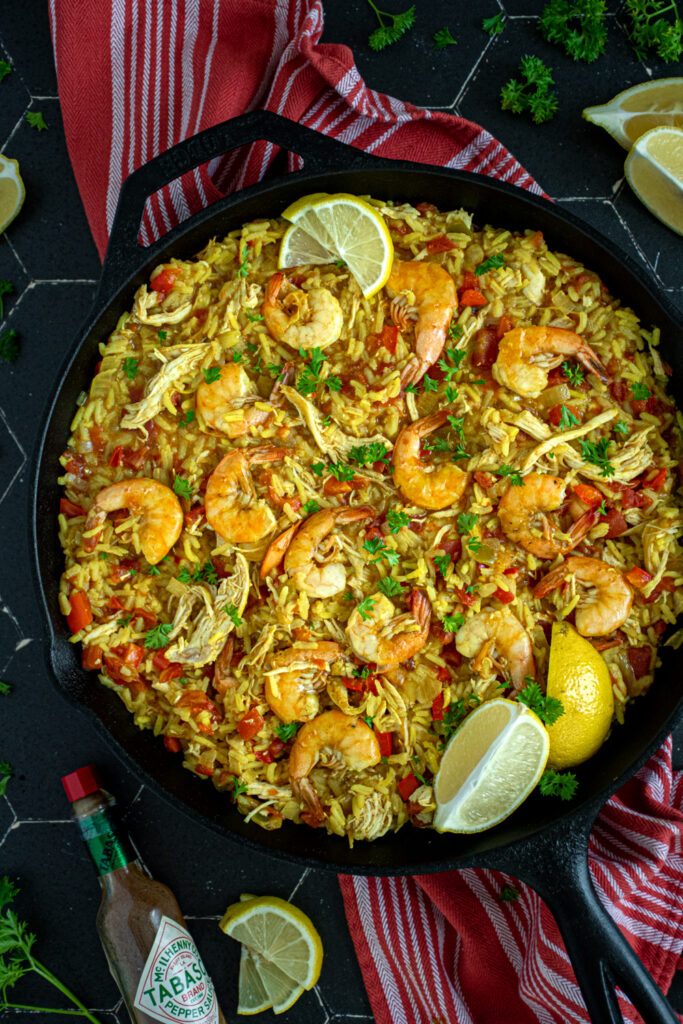 Close up of a pan filled with yellow chicken and shrimp paella with shrimp sitting on top with chunks of chicken, tomatoes, and bell peppers.
