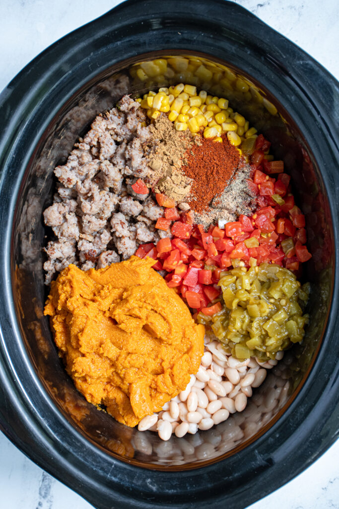 A black crockpot filled with the ingredients to make pumpkin chili, including maple sausage, white, beans, pumpkin, and tomatoes.