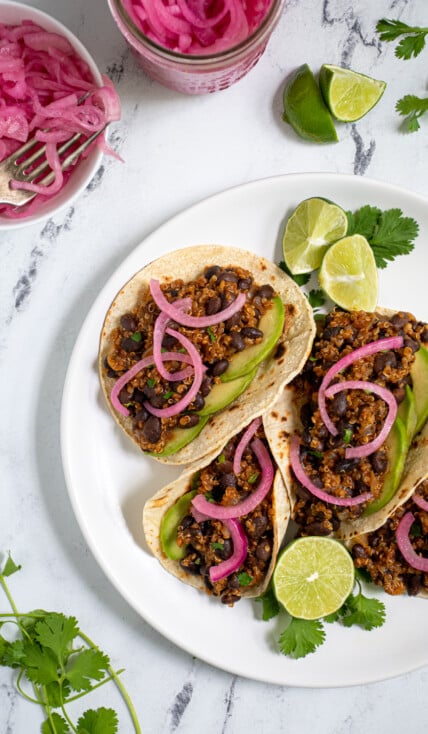 Overhead view of a white plate filled with black bean quinoa tacos garnished with cilantro, pickled onions, and avocado.