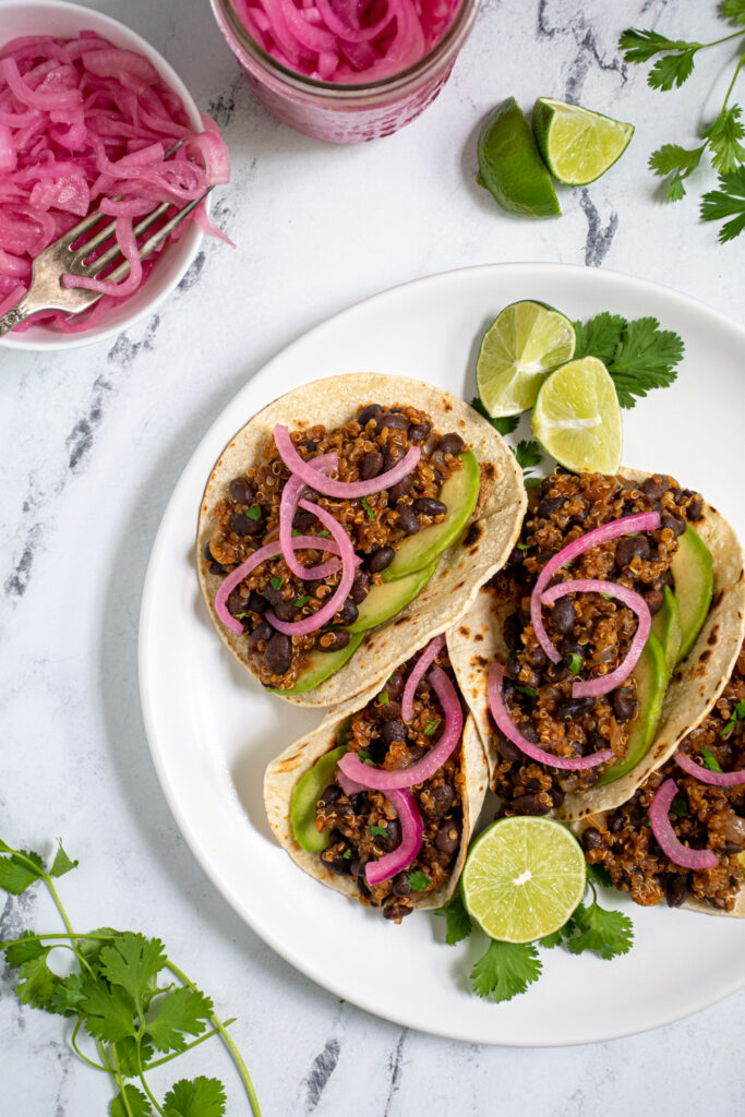 Overhead view of a white plate filled with black bean quinoa tacos garnished with cilantro, pickled onions, and avocado.