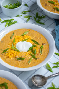 A white bowl of pumpkin chili on a white surface surround by more bowls and a lot of green onion garnishes.
