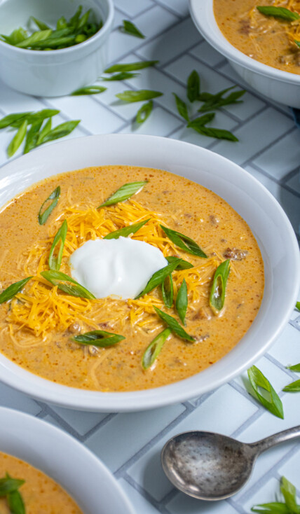 A white bowl of pumpkin chili on a white surface surround by more bowls and a lot of green onion garnishes.