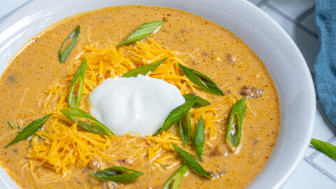 A white bowl of pumpkin chili on a white surface surround by more bowls and a lot of green onion garnishes.