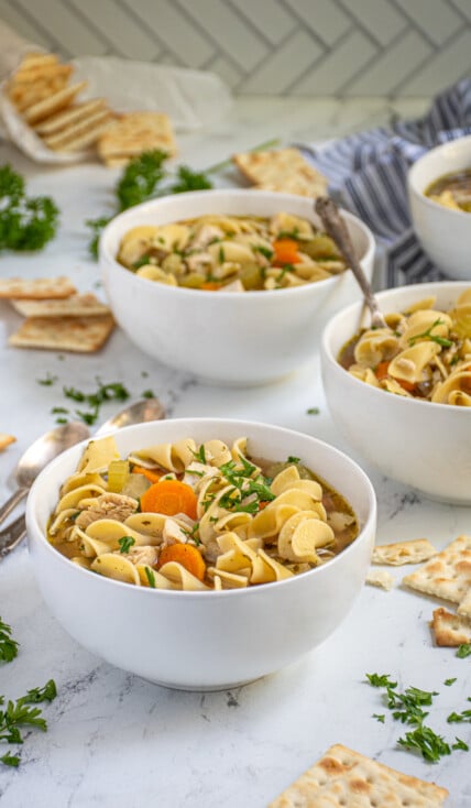 Countertop with four white bowls of easy chicken noodle soup showing off chewy egg noodles, big chunks of chicken, and vegetables garnished with fresh parsley.