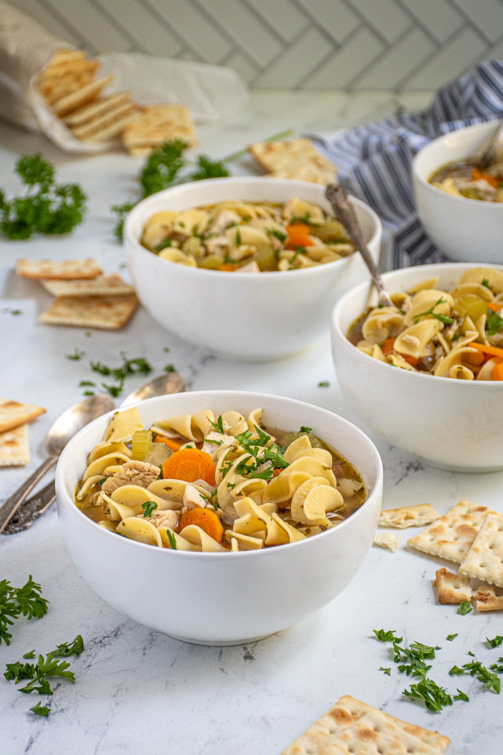 Countertop with four white bowls of easy chicken noodle soup showing off chewy egg noodles, big chunks of chicken, and vegetables garnished with fresh parsley.