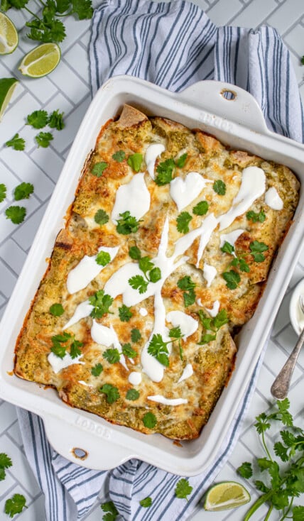 Overhead view of green chicken enchiladas in a white baking dish, topped with melty cheese, sour cream, and cilantro.