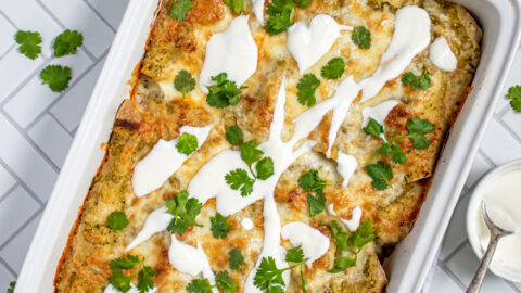 Overhead view of green chicken enchiladas in a white baking dish, topped with melty cheese, sour cream, and cilantro.