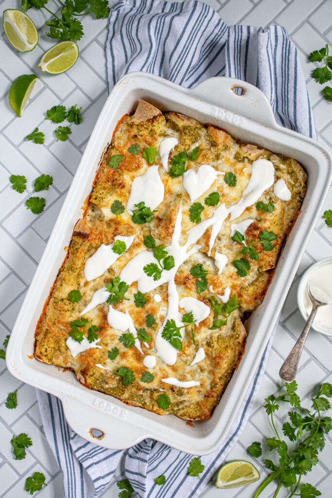 Overhead view of green chicken enchiladas in a white baking dish, topped with melty cheese, sour cream, and cilantro.