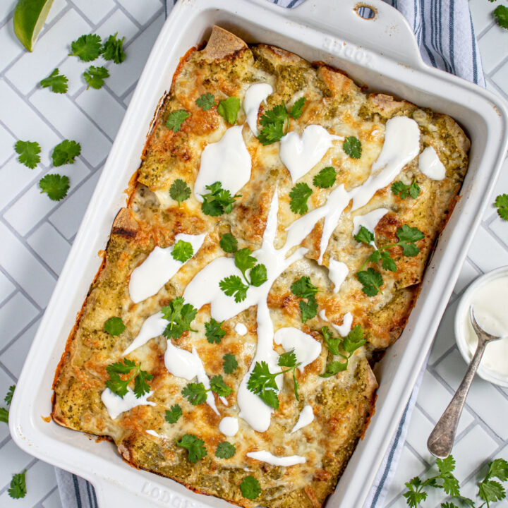 Overhead view of green chicken enchiladas in a white baking dish, topped with melty cheese, sour cream, and cilantro.