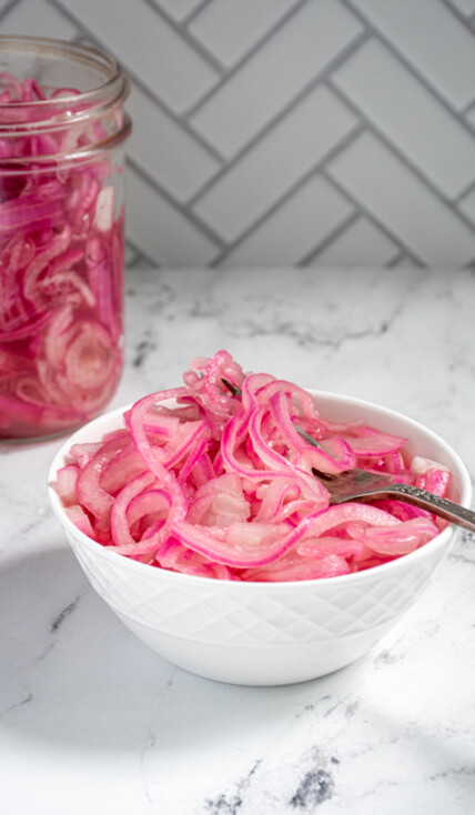 A white bowl with quick pickled red onions and a fork on a countertop next to a jar full of quick pickled onions.