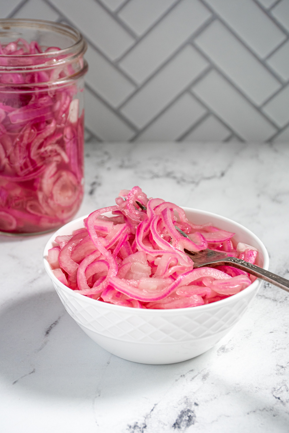 A white bowl with quick pickled red onions and a fork on a countertop next to a jar full of quick pickled onions.