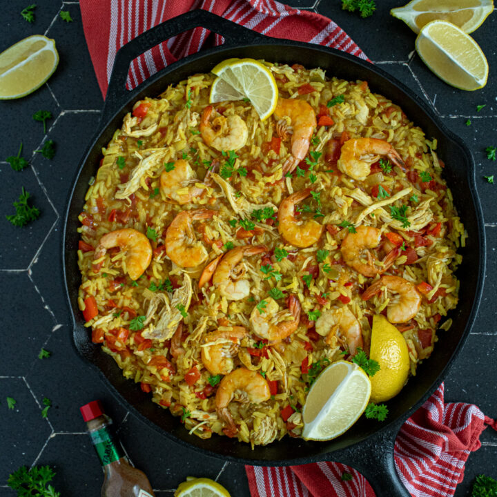 Overhead view of a black pan full of chicken and shrimp paella showing off yellow rice, chicken, red bell pepper, and cooked shrimp garnished with fresh parsley and lemon wedges.