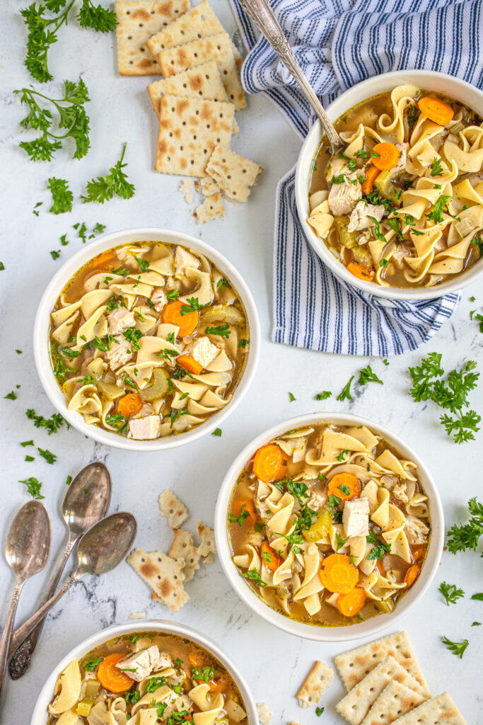 Flatlay photo of four white bowls of soup filled with easy chicken noodle soup with everything garnished with fresh parsley and served with saltine crackers.