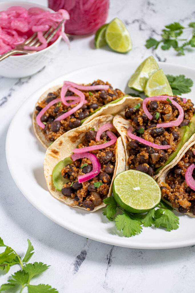 Plate with a bunch of black bean quinoa tacos in corn tortillas topped with pickled onions and avocado.