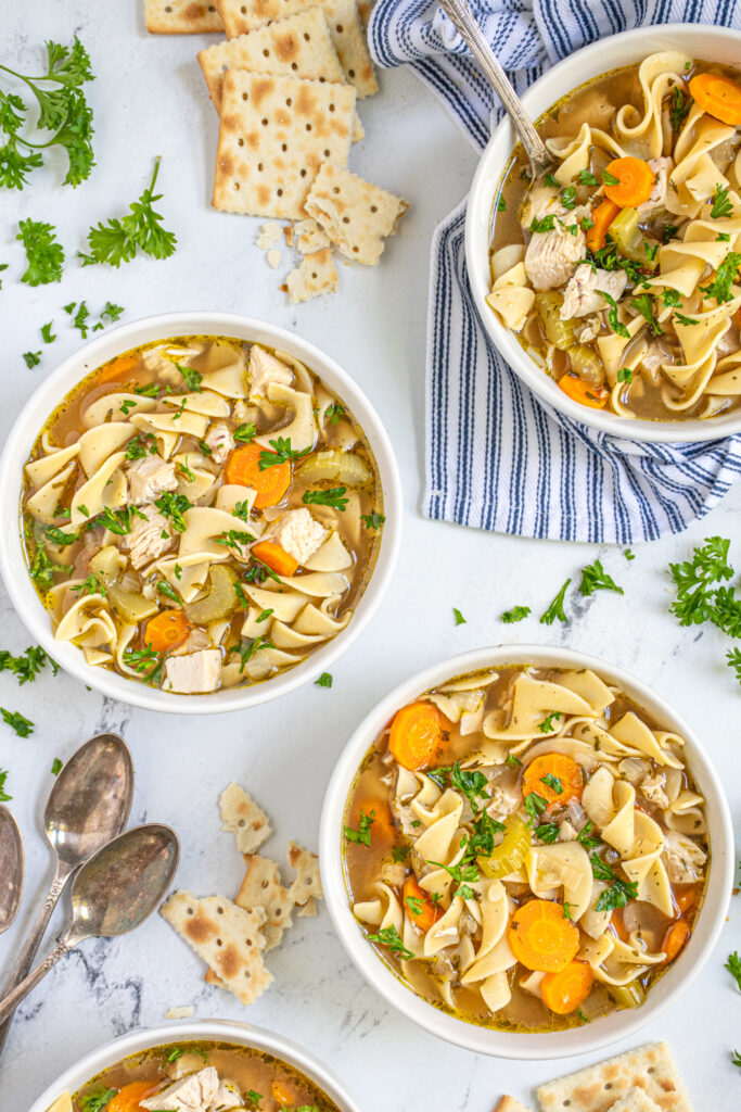 Overhead view of three white bowls filled with hearty stovetop chicken noodle soup garnished with fresh parsley and served with saltine crackers.