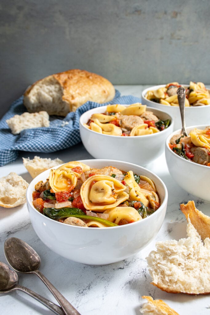 Tabletop with bowls of tortellini soup with chicken sausage and spinach surrounded by bread and spoons.