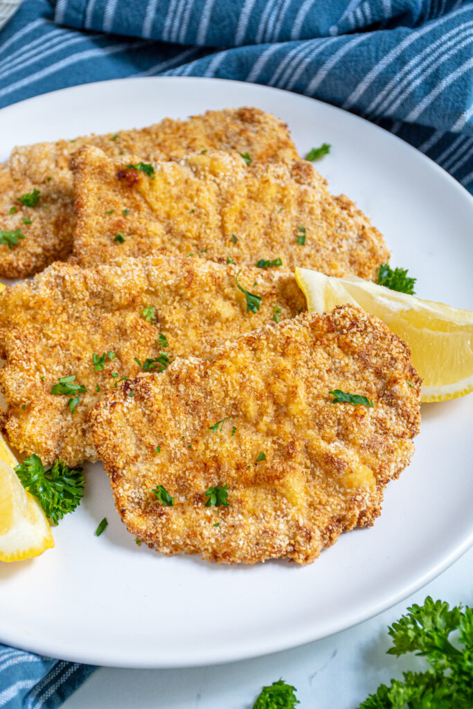 Close up of a row of air fryer pork schnitzels on a white plate with parsley and lemon wedges.
