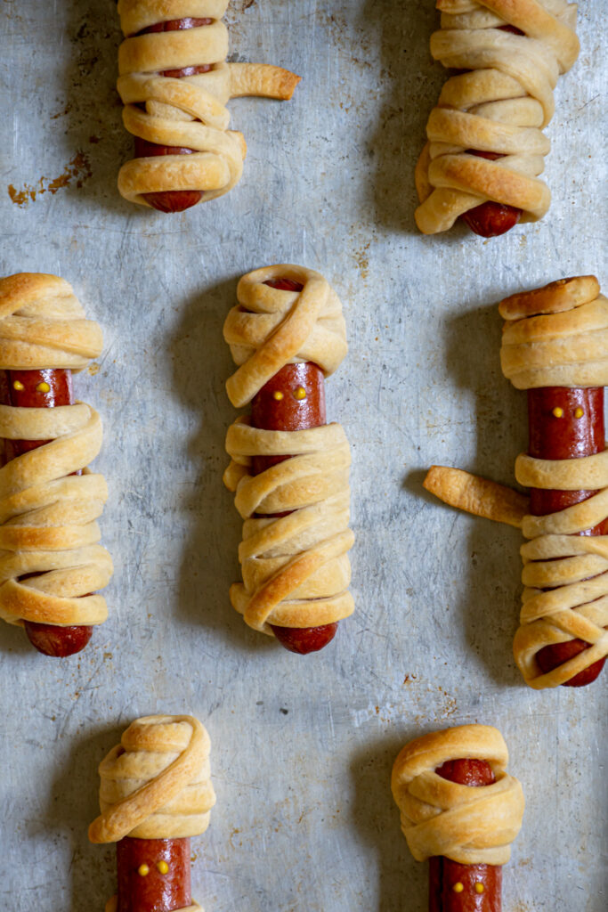 Close up of a mummy hot dogs on a sheet pan, wrapped in golden dough with mustard dot eyes.