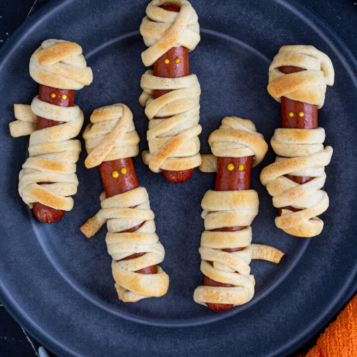 Close up of a black plate with 5 golden brown mummy dogs with mustard dot eyes.