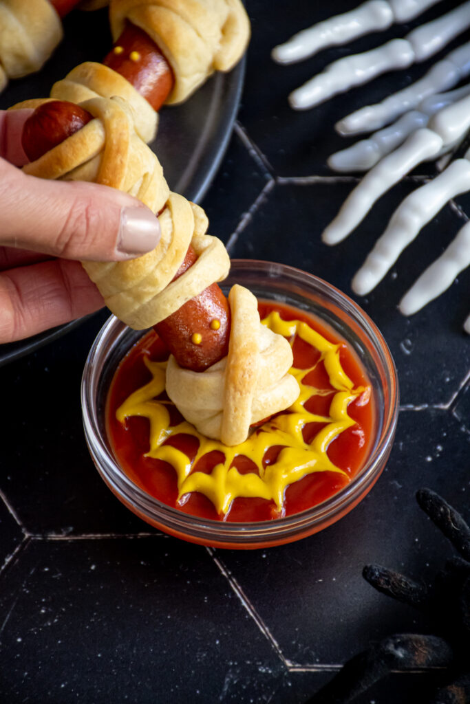 A mummy dog being dunked into a cup of ketchup and mustard that is in the design of a spider web.