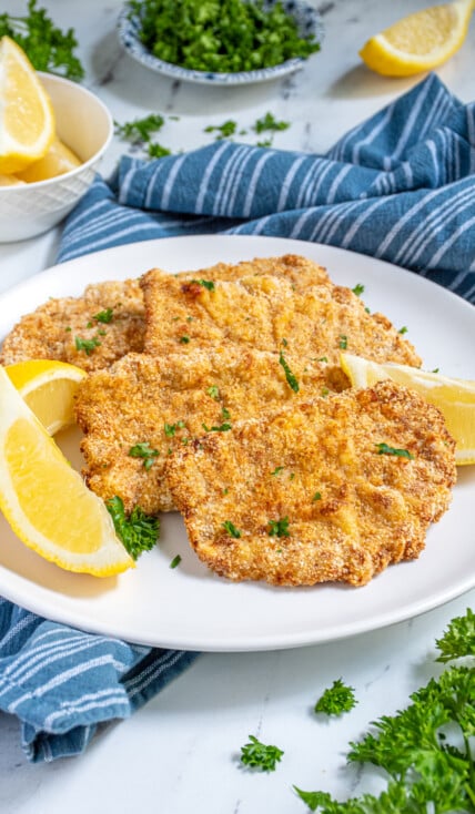 Air Fryer Pork Schnitzel lined up on a white plate garnished with fresh parsley and surrounded by lemon wedges.
