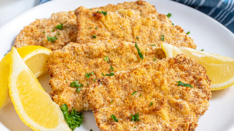 Air Fryer Pork Schnitzel lined up on a white plate garnished with fresh parsley and surrounded by lemon wedges.
