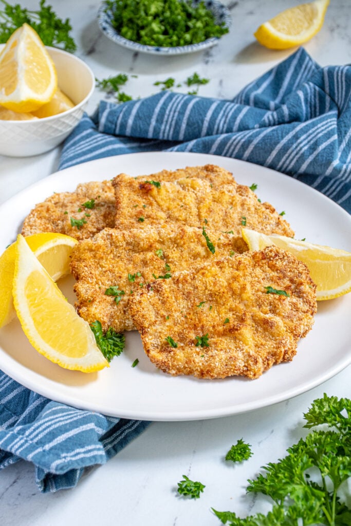 Air Fryer Pork Schnitzel lined up on a white plate garnished with fresh parsley and surrounded by lemon wedges.