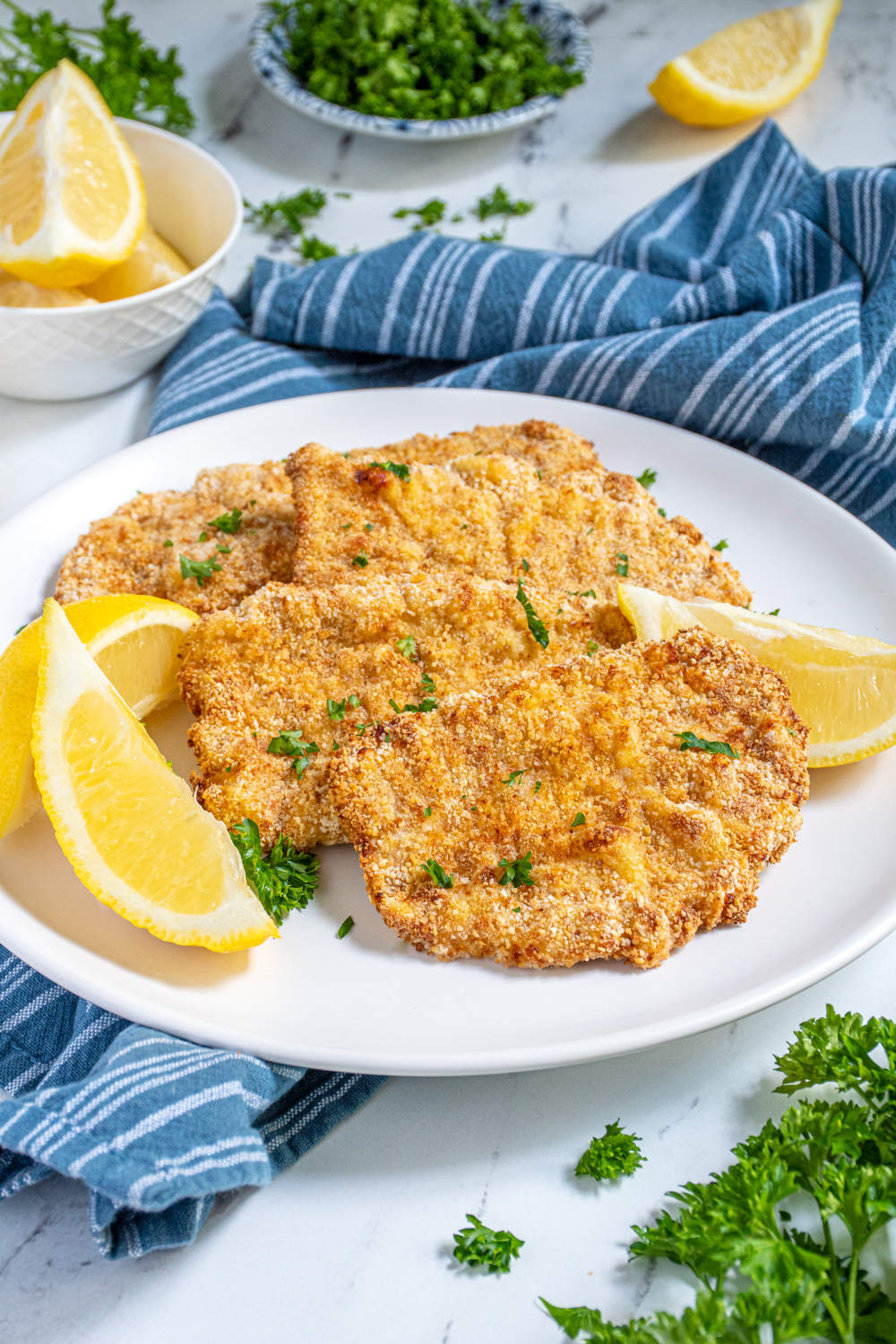 Air Fryer Pork Schnitzel lined up on a white plate garnished with fresh parsley and surrounded by lemon wedges.