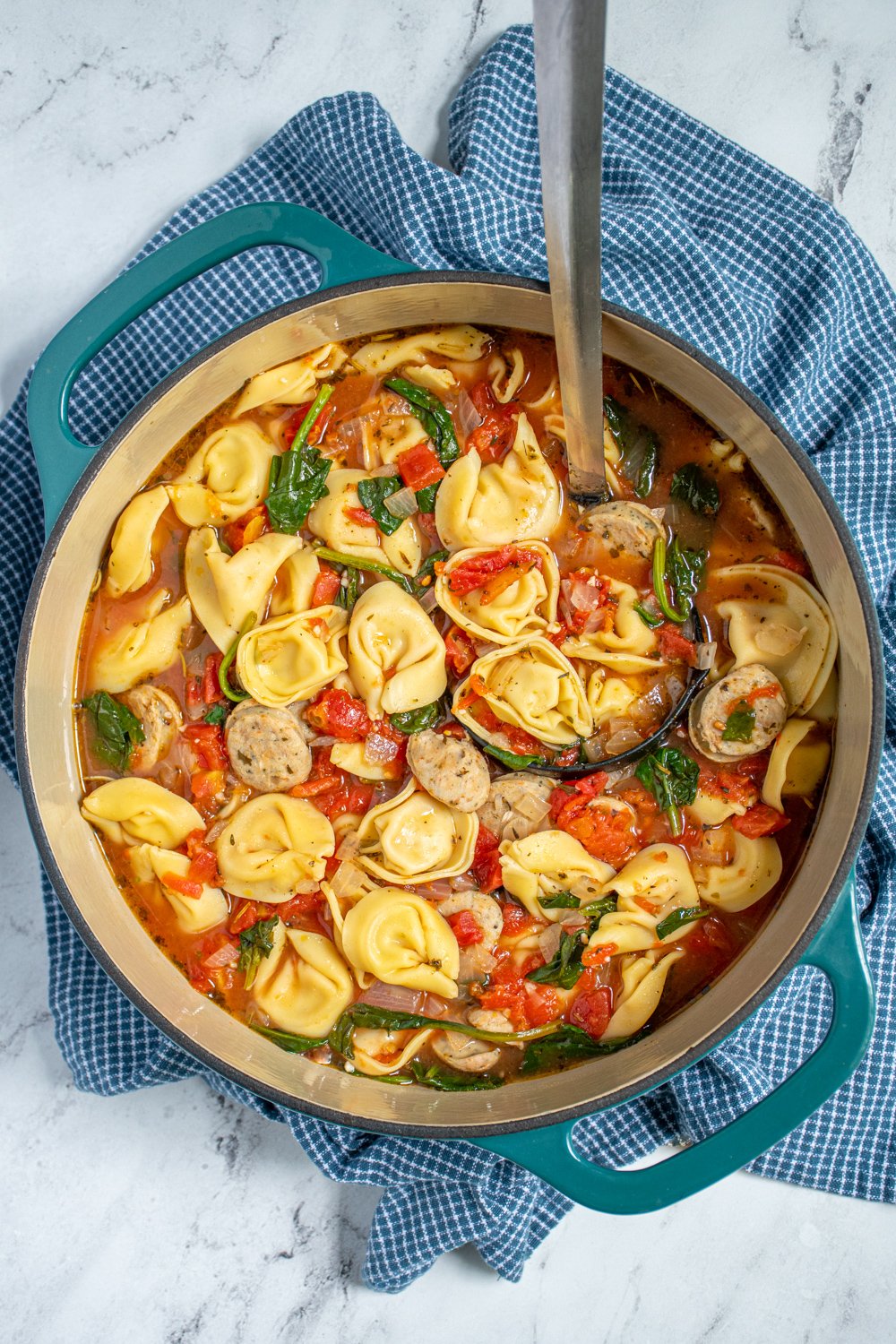 Overhead view of a large teal dutch oven full of chicken sausage tortellini soup with tomatoes and spinach.