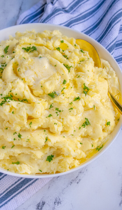 White bowl full of fluffy mashed potatoes dripping with melted butter and garnished with fresh parsley.