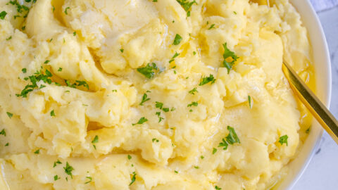 White bowl full of fluffy mashed potatoes dripping with melted butter and garnished with fresh parsley.