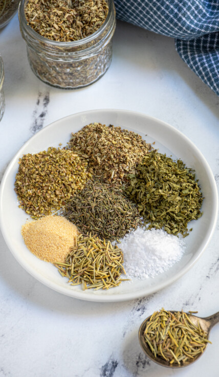 Small plate holding 7 little piles of different herbs and spices that make up Italian Seasoning.