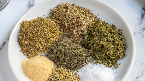 Small plate holding 7 little piles of different herbs and spices that make up Italian Seasoning.