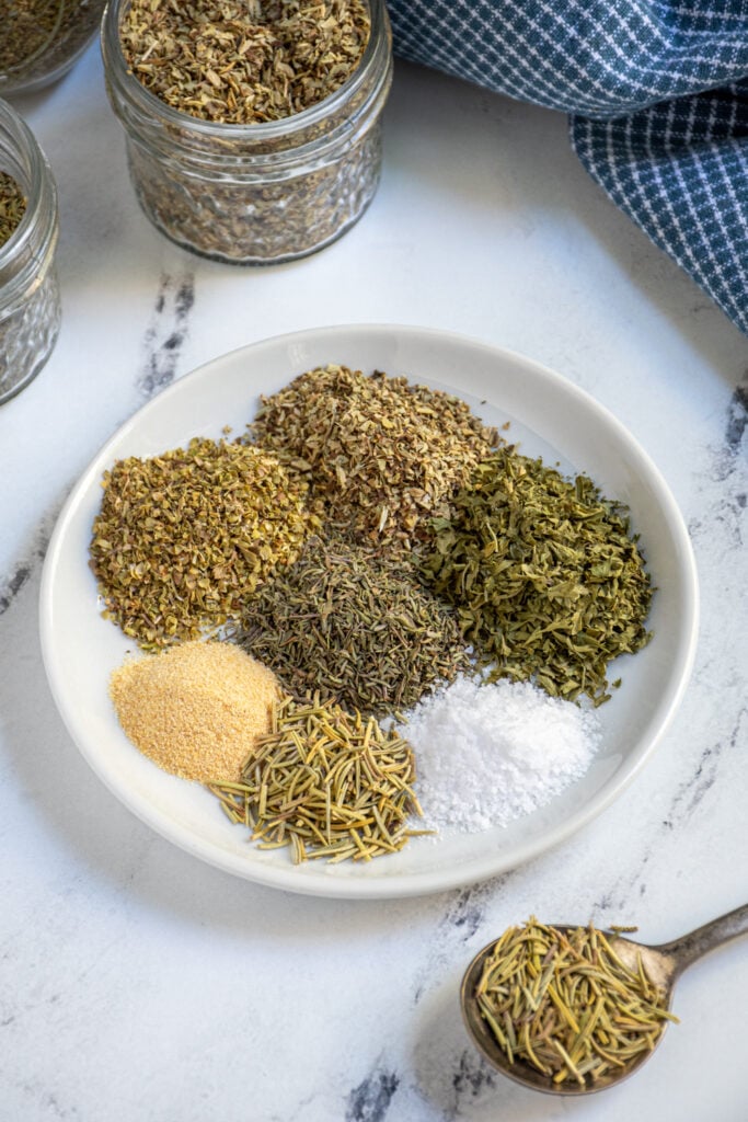 Small plate holding 7 little piles of different herbs and spices that make up Italian Seasoning.