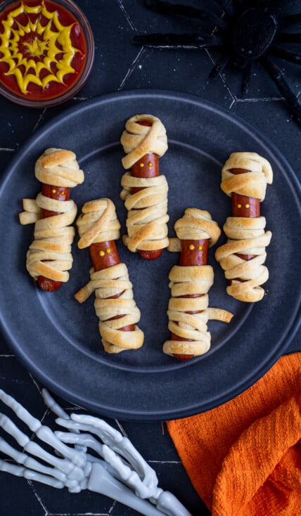On a black background on a black plate sits five mummy hot dogs wrapped in golden brown dough and mustard dot eyes.