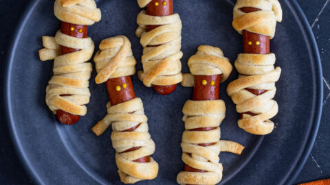 On a black background on a black plate sits five mummy hot dogs wrapped in golden brown dough and mustard dot eyes.