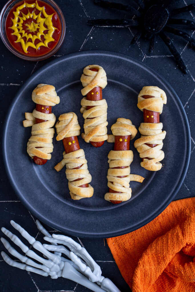 On a black background on a black plate sits five mummy hot dogs wrapped in golden brown dough and mustard dot eyes.
